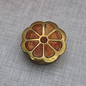 Pode incluir: Uma pequena caixa decorativa redonda com um design floral. A caixa tem uma moldura de metal dourado e um topo segmentado em forma de flor com secções castanho-avermelhadas e bege. A caixa está sobre uma superfície cinzenta clara, com flores cor-de-rosa e amarelas no fundo.