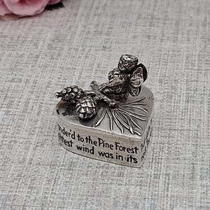 May include: A silver heart-shaped trinket box with a detailed relief design of pinecones and a bird. The box has an inscription that reads "der'd to the Pine Forest, west wind was in its". Decorative floral elements are in the background.