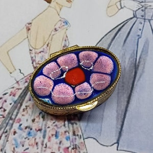 May include: An oval-shaped, vintage trinket box with a gold-colored frame. The lid features a floral design with pink and red accents on a blue background. The background includes a vintage fashion illustration.