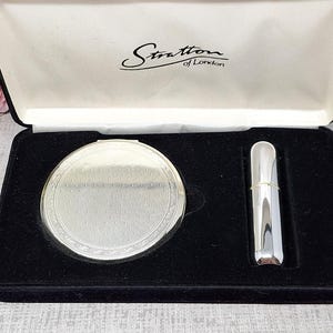 May include: A Stratton of London set in a black velvet-lined presentation box containing a round silver-tone compact mirror with a textured finish and a matching cylindrical silver-tone lipstick case, both nestled in a black foam insert.