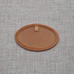 Puede incluir: Un parche ovalado marrón con un borde difuso y una pequeña pestaña en la parte superior. El parche está hecho de un material suave y texturizado. El fondo es neutro.