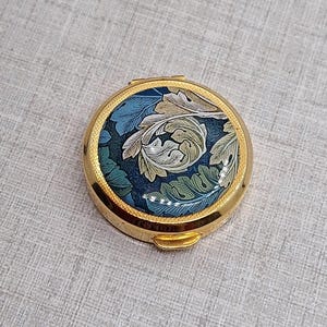 May include: A circular, gold-coloured pill box with a floral pattern. The lid displays a dark blue backdrop with a design of stylised leaves in green, cream, and blue. The box is closed and has a small clasp.
