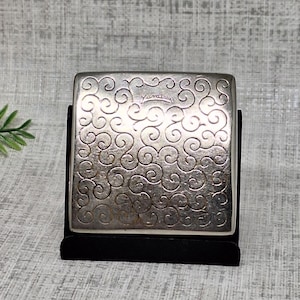 May include: A square silver-toned metal compact case featuring an embossed swirling scroll pattern across the front surface. The word YARAT is engraved in the center of the design. The compact is displayed upright on a small black stand.