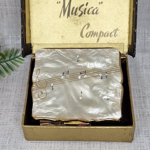 May include: A square Stratton Musica Compact powder case with a pearlescent white finish, featuring a gold-tone musical staff and notes on the lid. The case is displayed inside its original gold-colored box with the text Stratton Musica Compact.