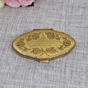 以下が含まれることがあります： 蓋にバラと葉のフローラル模様が刻印された、楕円形の真鍮製コンパクトケース。金属の表面はブラシ仕上げの質感で、ケースの縁はスカラップ状になっており、中央のデザインの周りに装飾的なフレームを作り出しています。