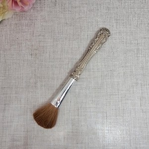 Op de afbeelding: Een zilverkleurige make-up borstel met een decoratief handvat en een bruine borstelkop. De borstel staat schuin op een lichtgrijze ondergrond, met roze en witte bloemen op de achtergrond.