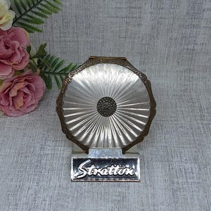 May include: A vintage Stratton compact mirror with a silver-tone, fan-shaped design and a dark center. The compact has a decorative, ornate frame and a rectangular base with the name "Stratton" in script.