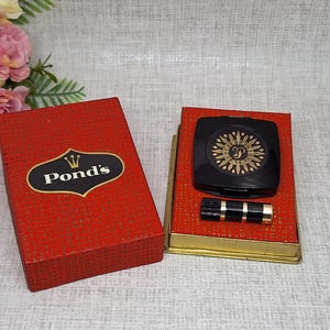 May include: A vintage red box with the Pond's logo next to a black compact with a gold design and a black and gold lipstick. The items are on a red and gold case. A bouquet of pink and yellow flowers is in the background.