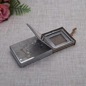 Op de afbeelding: Een rechthoekige, zilverkleurige thee-infuser met een scharnierend deksel en een fijnmazig filter. De infuser is gedeeltelijk open en toont het gaas. De doos heeft een geëtst ontwerp. Een kleine bloemstuk staat op de achtergrond.