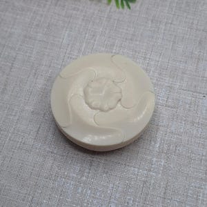 May include: A round, off-white decorative object with a floral design in the center and swirling patterns around it. The object is made of a solid material and sits on a textured surface. The design is raised, creating a three-dimensional effect.