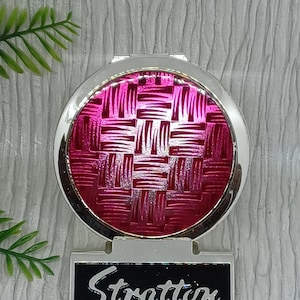 May include: A silver-toned compact mirror with a vibrant pink woven design on the front. The compact has a rectangular base with the name "Stratton" in cursive script. The background is a textured white surface.