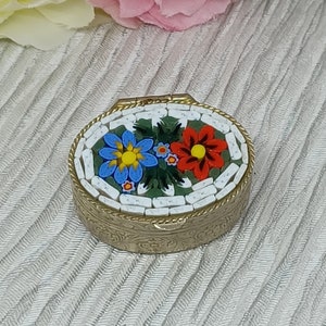 May include: An oval, gold-toned trinket box with a mosaic floral design on the lid. The mosaic features blue and red flowers with yellow centers on a green background. The box is set against a textured, light-colored fabric.