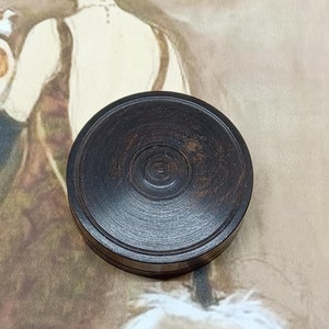 Könnte beinhalten: Eine dunkelbraune, runde Holzbox mit konzentrischen Kreisen auf dem Deckel. Die Box befindet sich vor einer Vintage-Illustration einer Frau in einem Kleid. Ideal zur Aufbewahrung kleiner Gegenstände.