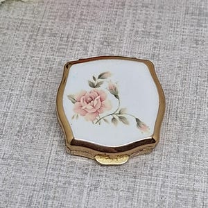 Puede incluir: Un pequeño espejo compacto con forma de escudo y marco de metal dorado. El centro presenta una incrustación de esmalte blanco decorada con una delicada ilustración de una rosa rosada con hojas verdes y un pequeño capullo en un tallo fino.