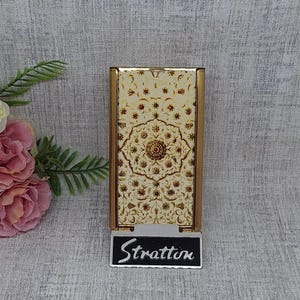 May include: A vintage Stratton compact mirror with a gold-tone frame and a cream-colored face featuring a detailed floral design. The compact has a rectangular shape and the name "Stratton" is displayed on a small black sign.