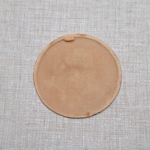 Puede incluir: Un parche circular de color tostado con una textura suave similar al ante y un borde cosido. El parche presenta una pequeña lengüeta elevada en el borde superior, ofreciendo un diseño limpio y sencillo para diversas aplicaciones textiles.