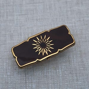 Könnte beinhalten: Eine rechteckige, dunkelbraune Haarspange mit goldfarbenem Rand und einem goldenen Sonnenstrahl-Design. Die Spange hat einen dekorativen, gewellten Rand, vor grauem Hintergrund.