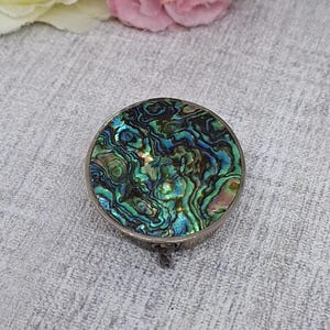 May include: A round, silver-toned pill box with an iridescent abalone shell top. The shell features swirling patterns of green, blue, and gold. The box is closed, and the hinge is visible.