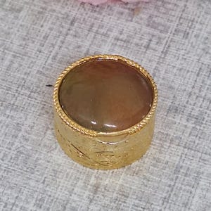 May include: A gold-coloured brooch featuring a round, multi-toned brown stone. The stone is encircled by a twisted rope-like border. The cylindrical base of the brooch has an embossed design. A classic accessory for adding a touch of elegance.