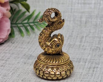 以下が含まれることがあります： 蓋に魚の彫刻が施された、小さな金色の金属製小物入れ。箱の底面には花びらや鱗のような模様が繰り返しあしらわれており、魚は曲線を描く体と細かな鱗、そして暗い色の目のようなアクセントが特徴的なデザインです。