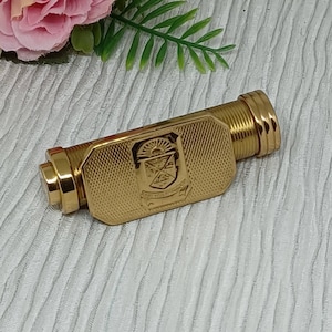 May include: A gold-colored, rectangular perfume bottle with a detailed crest design. The bottle has a ribbed, cylindrical end and a hinged cap. The background is a textured, white surface with pink flowers and green leaves.
