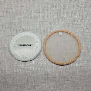 Puede incluir: Dos discos cosméticos circulares. Uno es una borla blanca con la palabra "manicare" impresa en texto negro en la superficie. El otro es un disco circular translúcido tipo malla con un borde de tela beige y una pequeña lengüeta blanca.