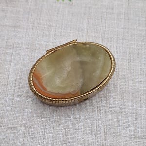 May include: An oval trinket box with a gold-coloured metal frame. The lid features a polished stone with shades of green, brown, and orange. The box is closed and rests on a light-coloured fabric surface.