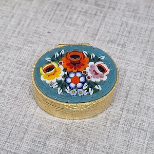 Könnte beinhalten: Eine kleine, runde, goldfarbene Pillendose mit einem floralen Mosaikdesign auf dem Deckel. Das Mosaik zeigt einen türkisfarbenen Hintergrund mit bunten Blumen in Orange-, Gelb- und Rosatönen. Die Dose ist geschlossen.