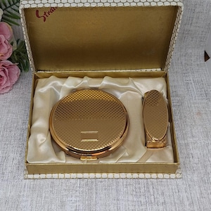 Könnte beinhalten: Ein Vintage-Set aus goldfarbenem Stratton-Puderetui und Lippenstifthalter in einer dekorativen Box. Das Puderetui und der Lippenstifthalter haben ein strukturiertes, geometrisches Muster. Die Box ist mit Satin gefüttert und hat einen gemusterten Deckel.
