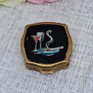 May include: A small, vintage, gold-toned compact with a black top featuring a cocktail and cigarette design. The compact has a rectangular shape with rounded edges. The design includes two cocktail glasses, a lit cigarette, and a blue ashtray.