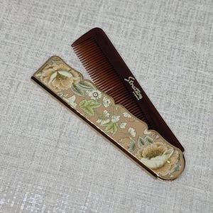 May include: A folding pocket comb with a dark brown plastic tooth section and a gold-tone metal casing. The exterior features a floral pattern with cream, tan, and green flowers on a light background. The word Stratton is printed on the comb.