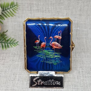 May include: A square Stratton compact case with a scalloped gold-tone metal edge. The front features a deep blue enamel background with a sunburst pattern and a painted scene of four pink flamingos standing in green water. The word Stratton is printed.