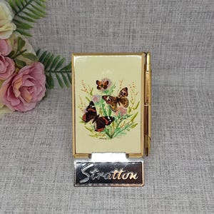 May include: A vintage Stratton compact mirror with a gold-tone frame. The front features a cream-colored background with a butterfly and floral design. The compact has a hinged opening and a silver-tone Stratton nameplate.