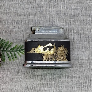May include: Vintage metal lighter with a black body and silver accents. The black panel features a gold-colored relief of a Japanese landscape with a mountain and buildings. The lighter has a flip-top ignition.