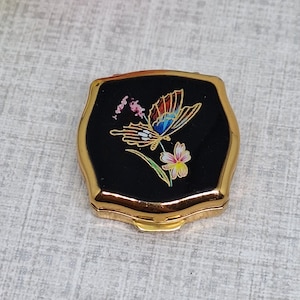 May include: A small, gold-toned metal compact case with a black enamel lid. The lid features a colorful graphic of a butterfly with orange, blue, and yellow wings perched on a small pink and white flower with green leaves and a stem.