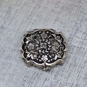 May include: A silver-toned, antique-style jewelry box with a floral design. The box is square with rounded edges and features intricate embossed flowers and leaves. The background is a light gray fabric.