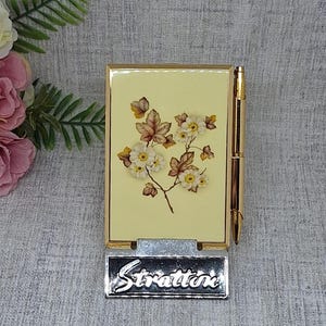 May include: A vintage Stratton compact with a floral design on a pale yellow background. The compact has a gold-tone frame and a matching pen. A silver Stratton nameplate is at the base.