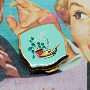 Cenicero de bolsillo Stratton ~ Esmalte azul/verde con un cigarrillo humeante en un cenicero en la parte superior junto a una planta de hiedra en maceta ~ Vintage de mediados de siglo