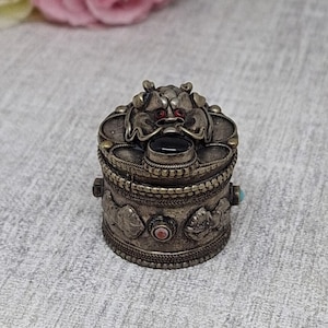 May include: A small, antique silver-colored box with a detailed lid featuring a stylized animal face with red eyes and a black stone. The box has decorative carvings and small, colorful stone accents.