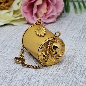 May include: A gold-colored, cylindrical container with a seashell design and a chain. The container has a decorative, round end with a skull and floral design. The chain is attached to a clasp.