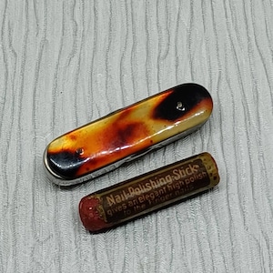 Op de afbeelding: Een vintage nagelpolijststokje met een rood en wit etiket waarop staat "Nail Polishing Sticks gives an elegant high polish to the finger nails". Het stokje ligt naast een klein nagelvijl met een schildpadpatroon.