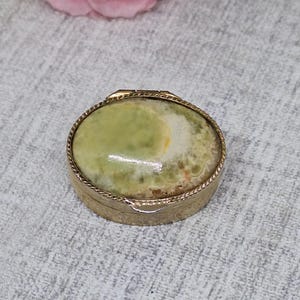 May include: A small, round, antique trinket box with a gold-coloured metal frame. The lid features an oval-shaped stone with shades of green, yellow, and white. The box is likely used for storing small jewellery items or keepsakes.