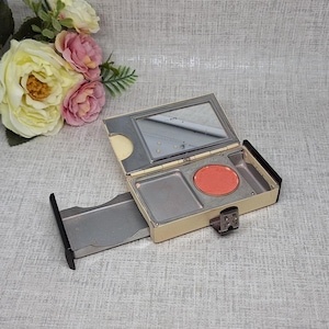 Op de afbeelding: Een rechthoekige, crèmekleurige compacte houder met een uitschuifbare lade en een scharnierend deksel. De binnenkant bevat een rechthoekige spiegel, een rond bakje met koraalkleurige blush en een leeg rechthoekig compartiment.