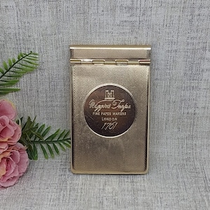 Könnte beinhalten: Ein goldfarbenes rechteckiges Etui mit strukturierter Oberfläche und Klappdeckel. Das Etui hat ein rundes Emblem mit dem Text "Wiggins Teape Fine Paper Makers London 1761". Das Etui wird auf einer hellgrauen Oberfläche mit floralen Akzenten präsentiert.