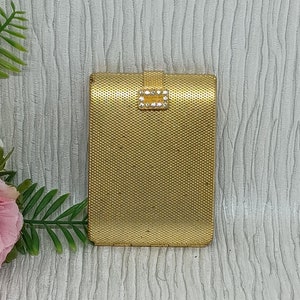 May include: A gold metal compact with a diamond-shaped clasp. The compact has a textured surface.