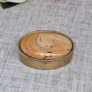 May include: A vintage pillbox with an oval, light brown stone lid set in a gold-coloured metal frame. The base of the box has an engraved pattern. A classic jewellery box, perfect for small items.