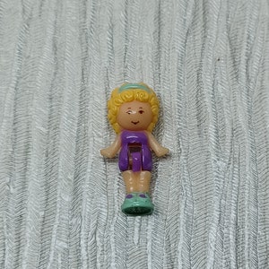 Puede incluir: Una pequeña muñeca de plástico con cabello rubio, un vestido morado y zapatos verdes. La muñeca es un juguete para niños.