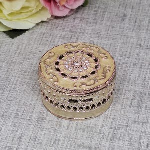 May include: A small, round jewelry box with a cream-colored lid and a decorative, openwork design on the sides. The lid features an ornate pattern with a central cluster of sparkling rhinestones. The box is likely made of metal.