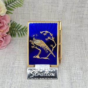 May include: A gold-toned compact with a blue enamel background featuring a detailed bird design. The compact has a silver Stratton nameplate. The bird is perched on a branch with gold-toned leaves.