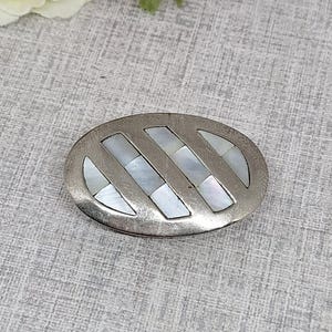 May include: Oval silver-toned belt buckle with a diagonal striped design. The stripes are inlaid with rectangular pieces of iridescent mother-of-pearl. The buckle has a vintage, antique appearance.